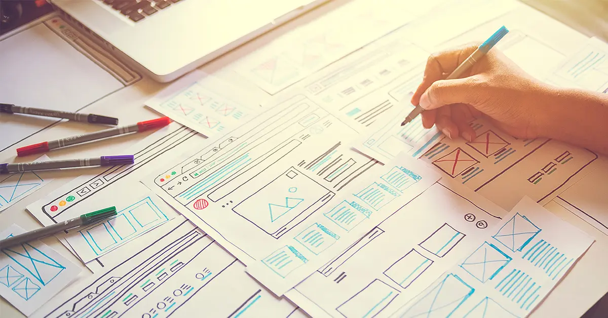 A person sketches website design wireframes on multiple sheets of paper spread across a desk. There are colored pens scattered around, and a laptop is partially visible in the background. The scene is lit with natural sunlight.