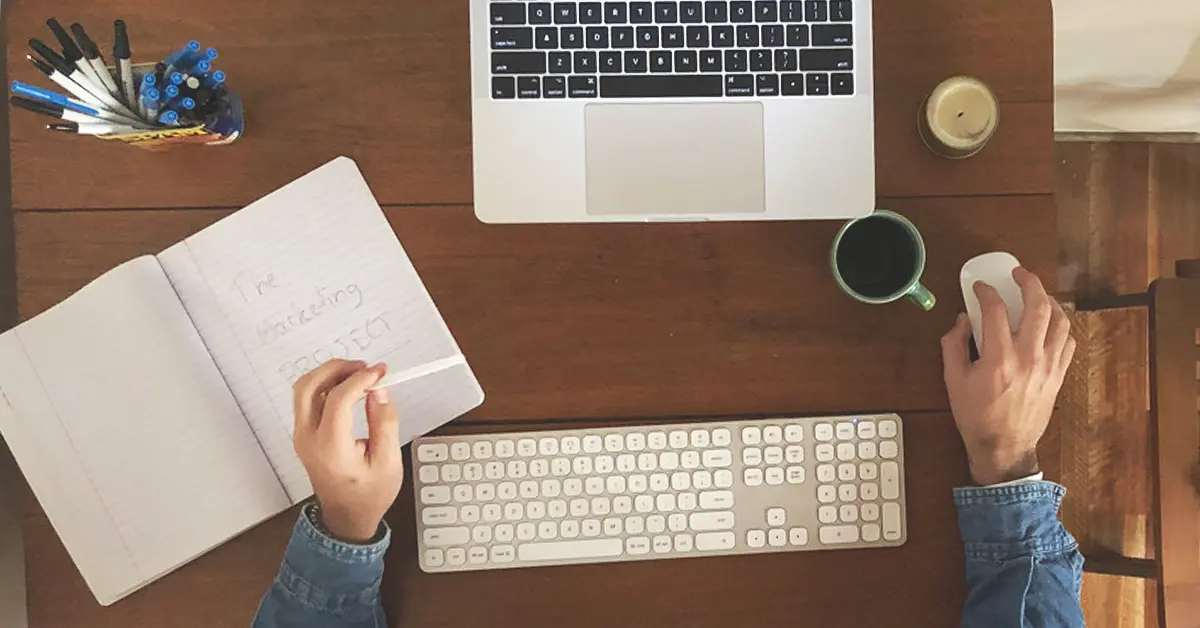 top view of a person's hand working on a laptop with wireless keyboard and with notes and bunch of pens and mug of coffee
