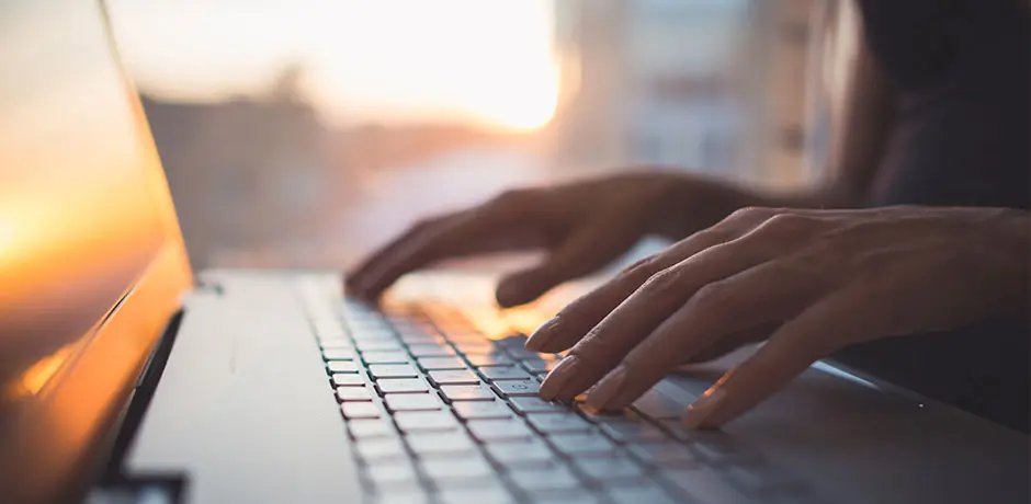 A person typing on a laptop keyboard with a blurred sunset in the background.