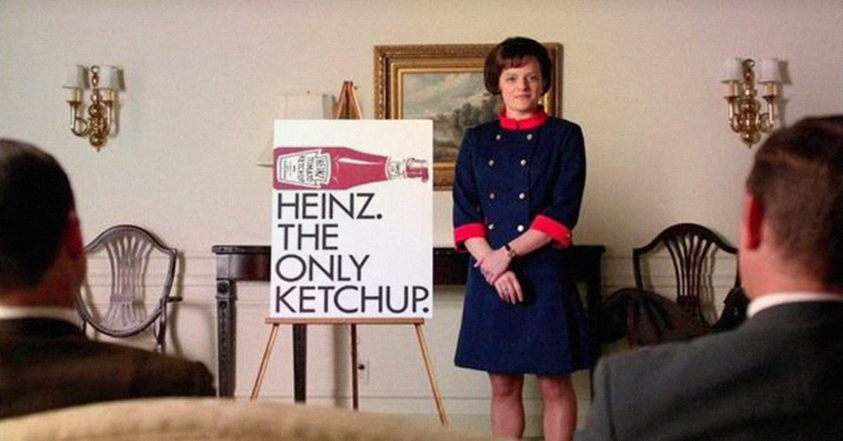 A woman in a blue dress with red accents stands next to a display featuring a Heinz ketchup bottle and the text "HEINZ. THE ONLY KETCHUP." She is in a room with beige walls, traditional-style chairs, and a framed painting in the background. Two people are seated in front of her, their backs partially visible.